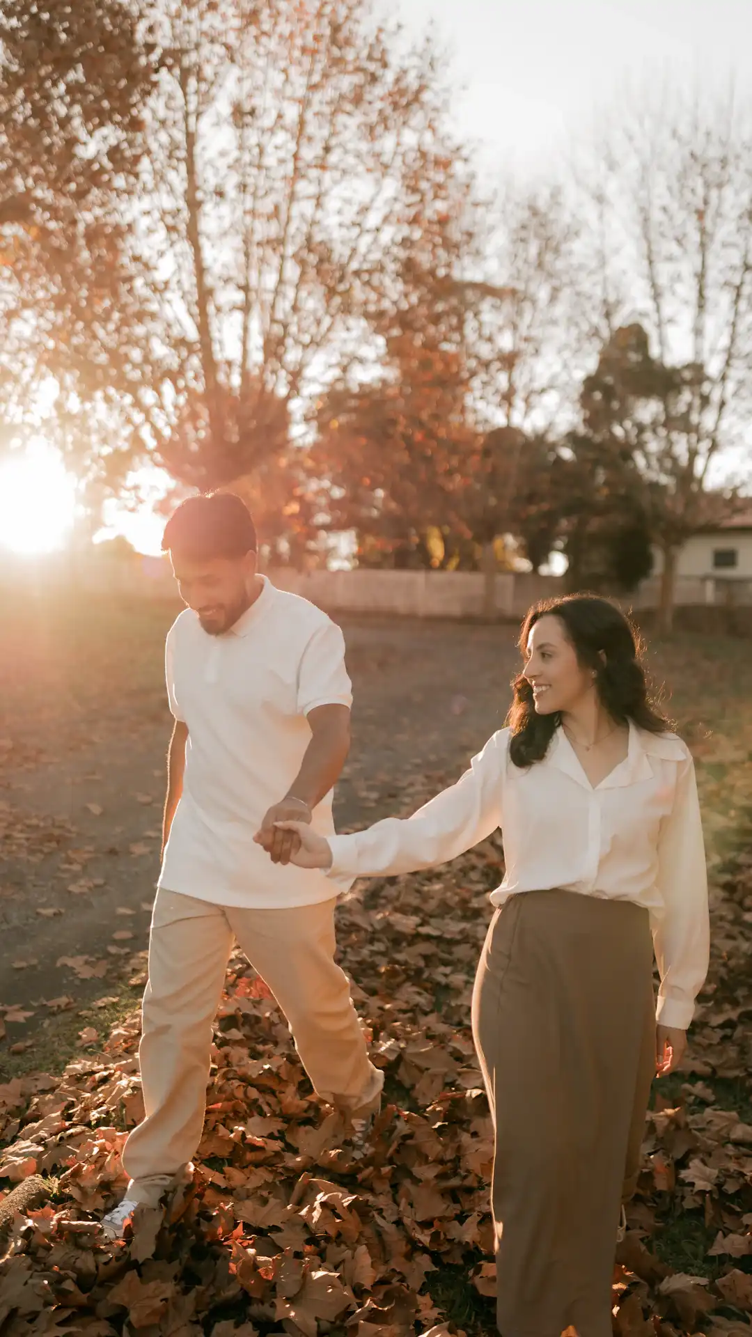 A couple holding hands walks through autumn leaves at sunset.