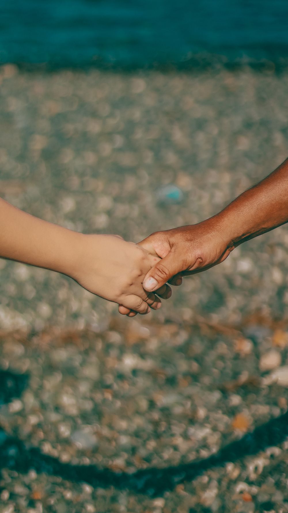 Close-up of two hands holding on a pebbly beach, symbolizing connection.