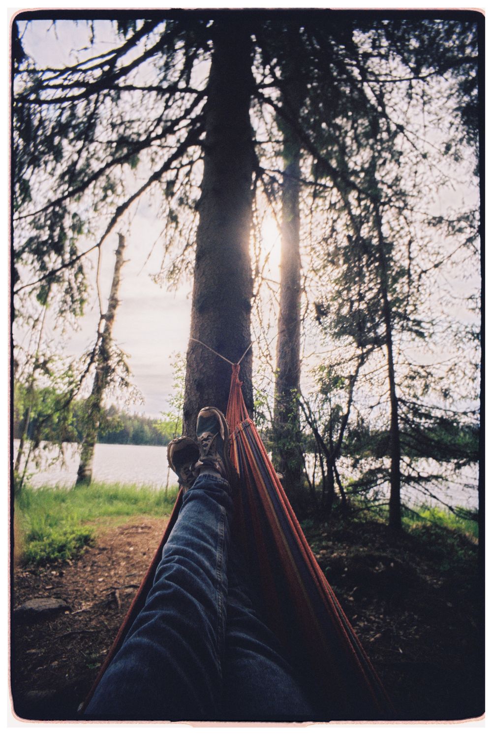 Person relaxing in hammock under trees by a lake at sunset, enjoying nature.