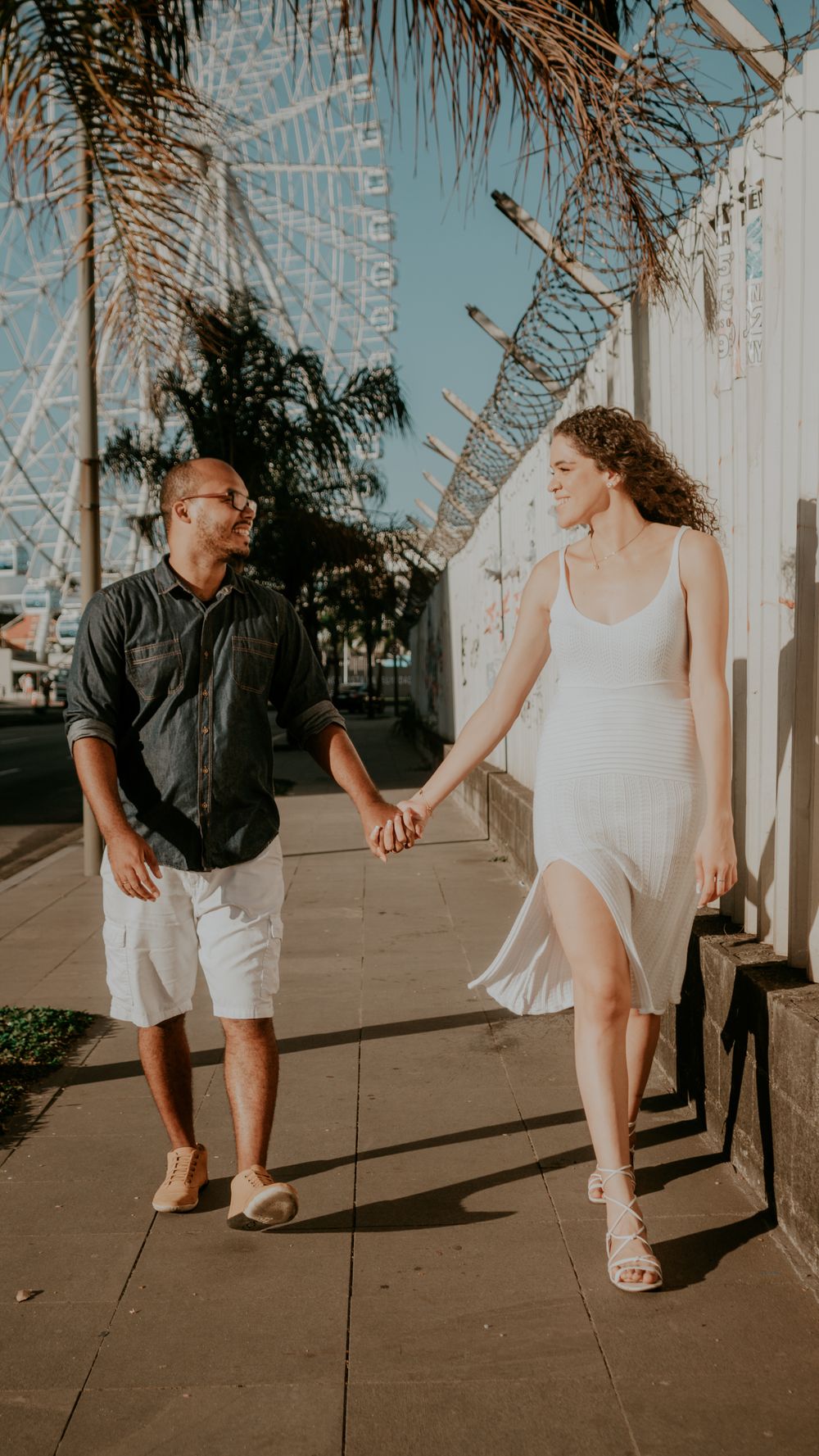 A loving couple strolls hand in hand along a sunny sidewalk near a Ferris wheel.